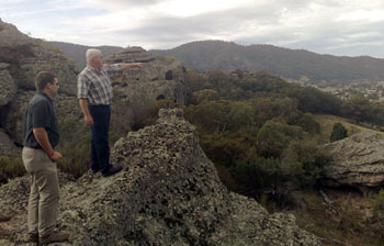 Standing on large rocks overlooking a green and rocky expanse.  Older mand with grey hair on higher rock pointing something out in the distance to younger man with blue shirt on lower rock.